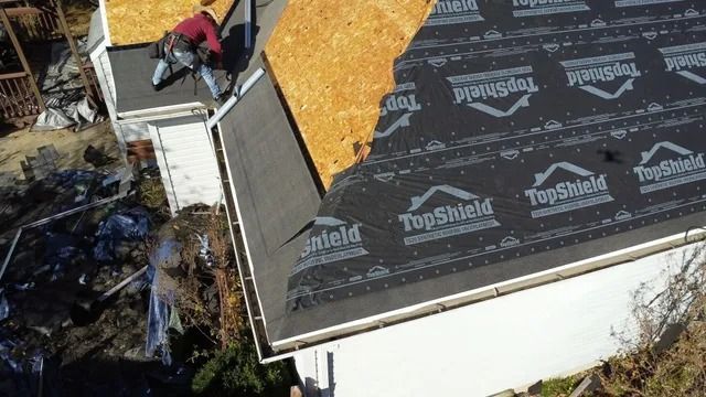 Aerial view of roofers installing shingles on a house roof with exposed plywood and black underlayment.