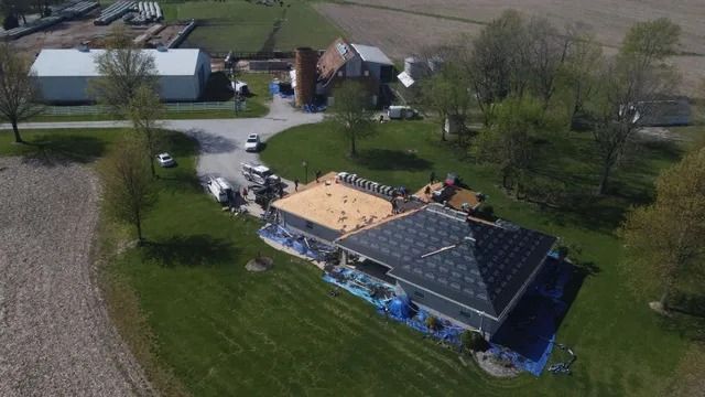 Aerial view of a house roof being replaced, with workers and materials on a grassy rural property.
