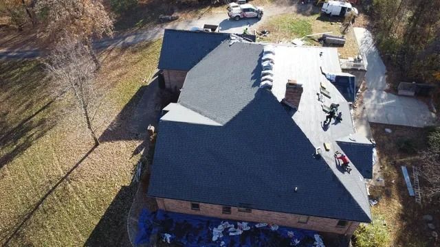Aerial view of a dark-roofed house with a white driveway and parked vehicles beside a wooded area