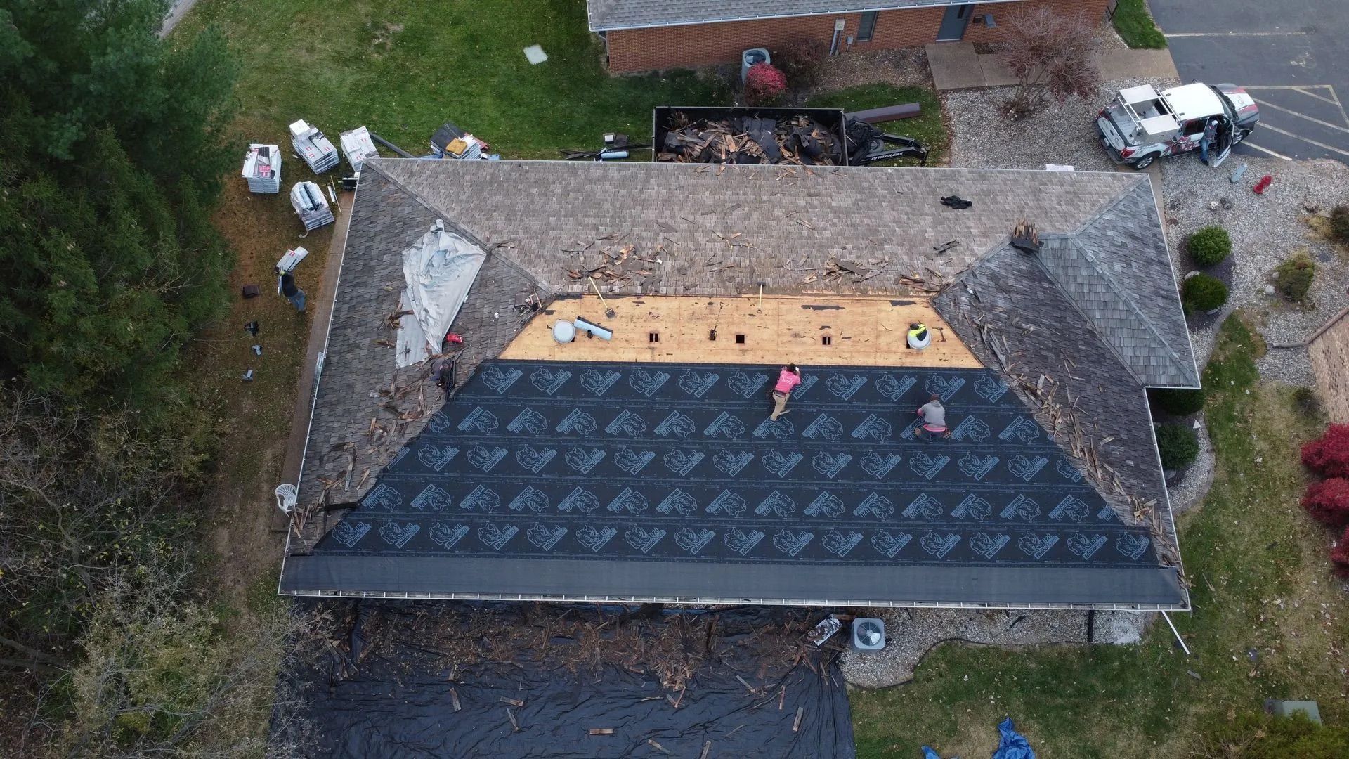 Aerial view of a roof under construction with dark shingles and workers on a partly covered section.