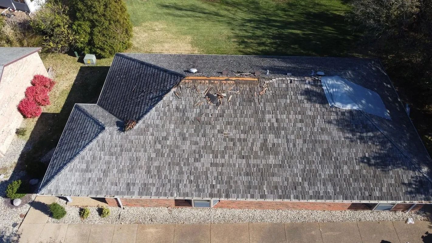 Aerial view of a house roof with dark shingles and a small black-roofed section beside green lawn.