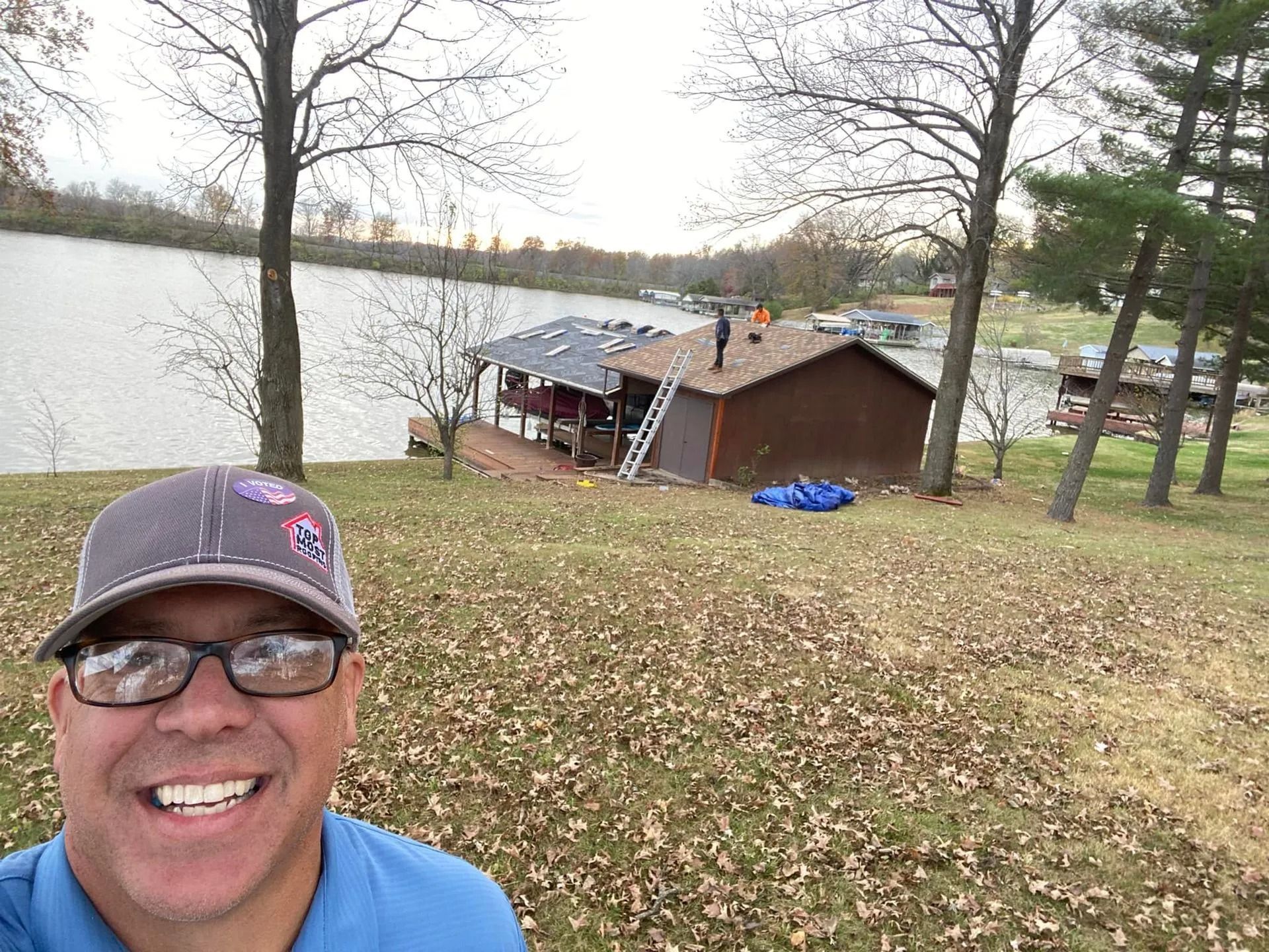 Smiling man in glasses and cap taking a selfie near a lakeside cabin in autumn