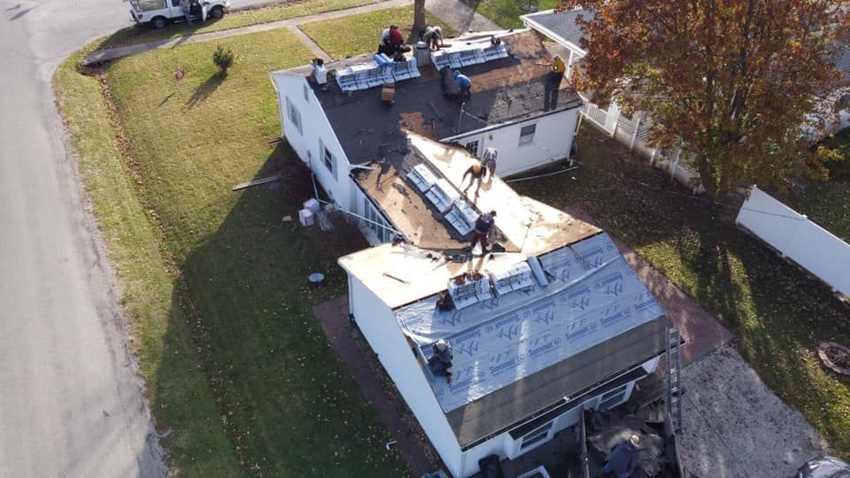 Aerial view of a damaged house roof with a large tarp, debris, and workers inspecting the damage.