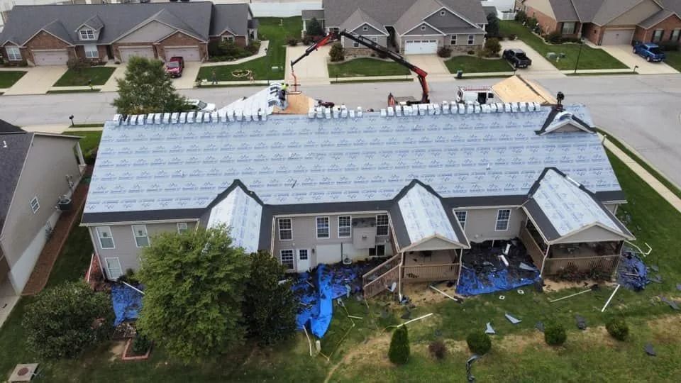 Aerial view of a long gray-roofed house with dormer windows and blue tarps in the yard, in a suburban neighborhood