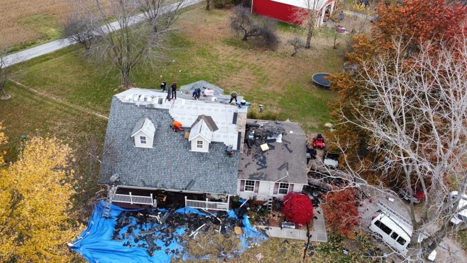 Aerial view of a damaged house with a collapsed roof, blue tarps, and debris in a yard.