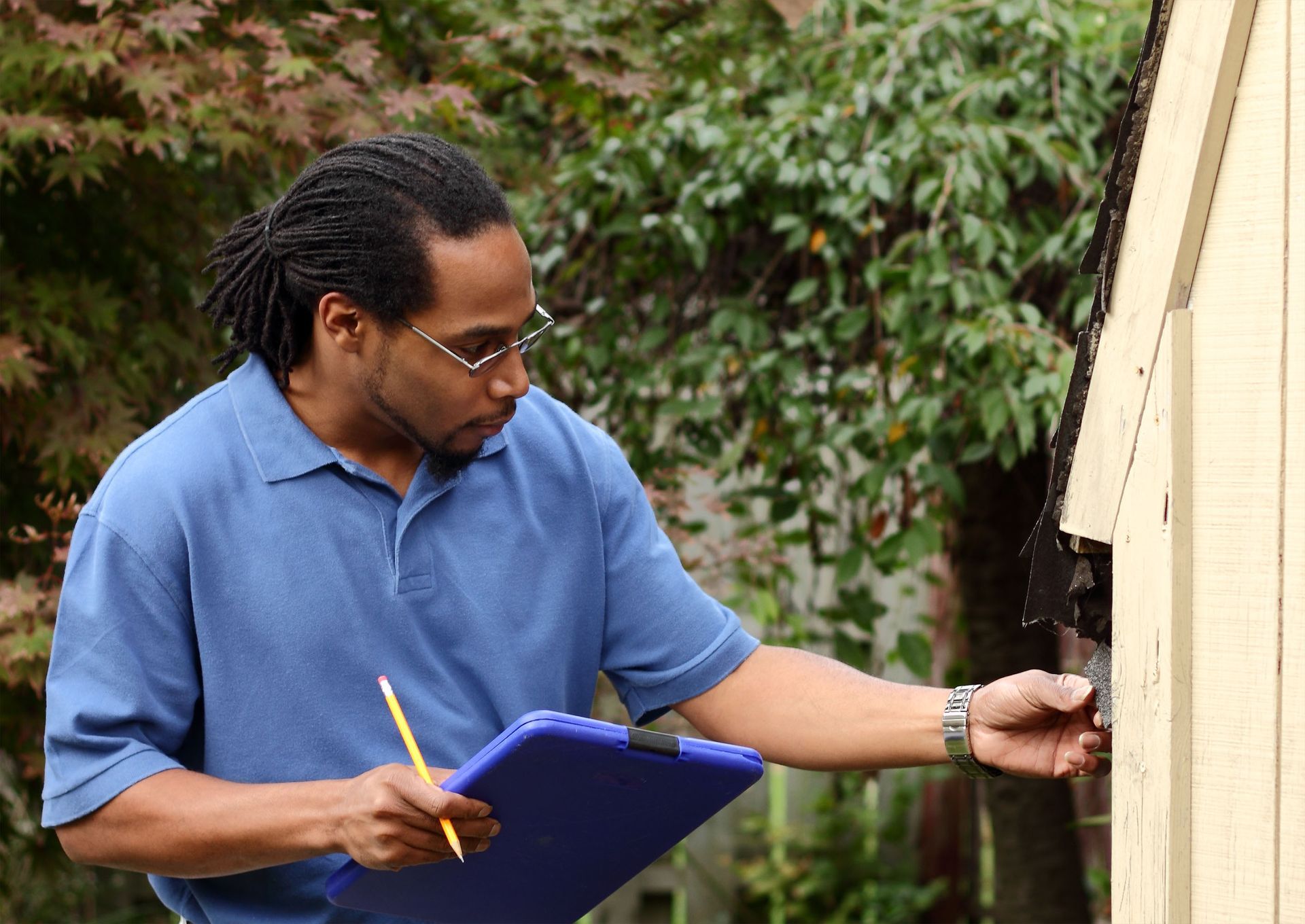 Person in a blue shirt writing on a clipboard while inspecting the side of a building outdoors