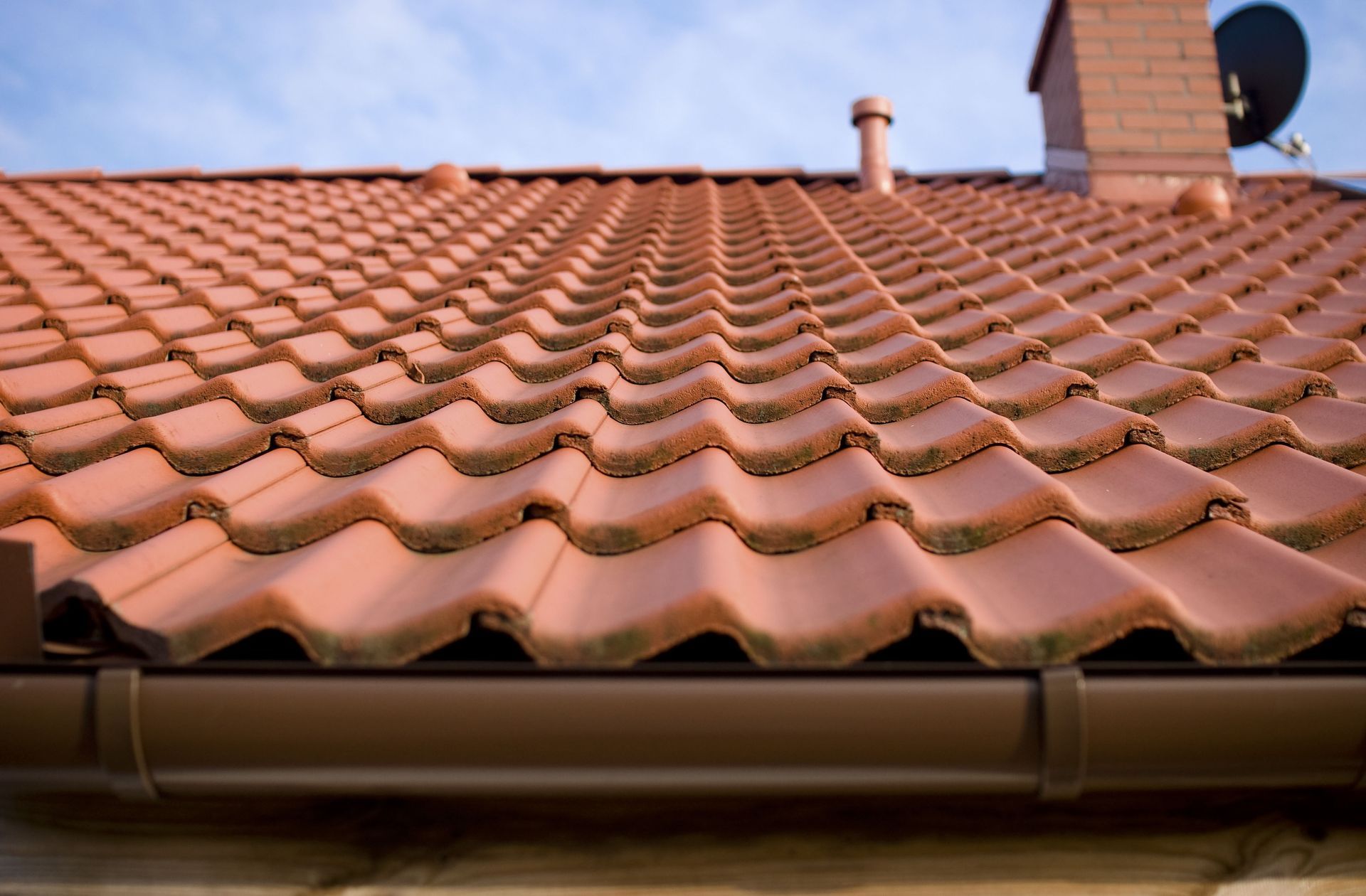 Red clay tile roof with chimney and gutter against a blue sky