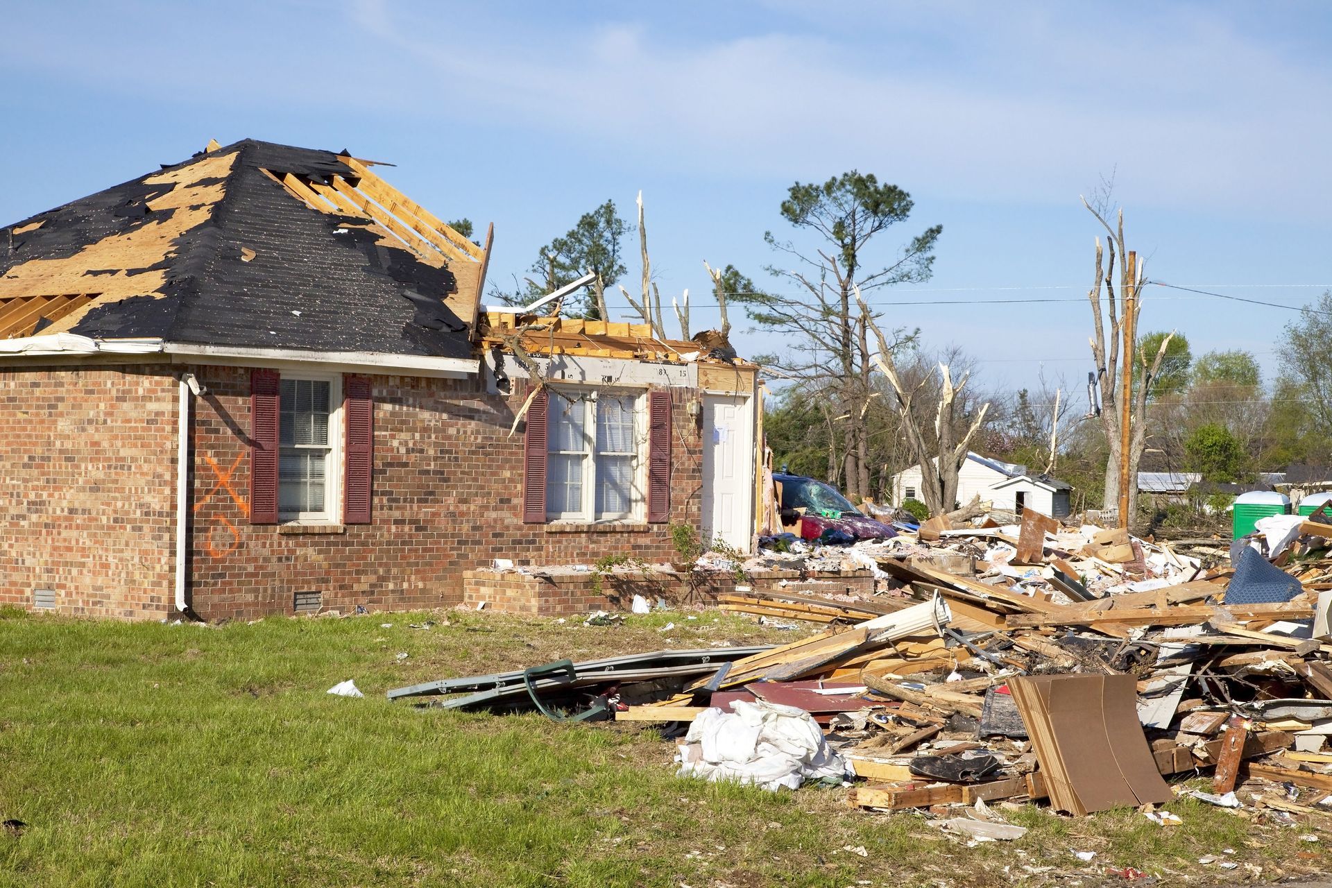 Damaged brick house with torn roof and debris scattered across a yard after a storm