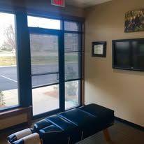 Chiropractor's office interior: window with blinds, treatment table, mounted TV, and artwork on wall.