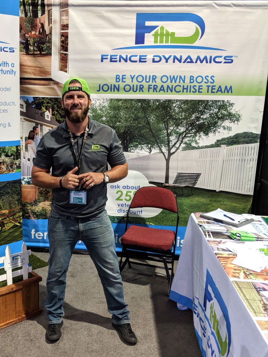 Man posing at a Fence Dynamics trade show booth with branded banner, tables, and chairs