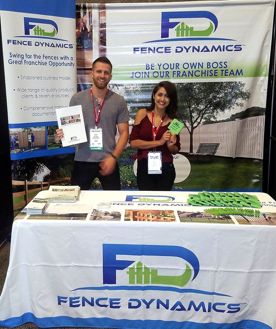 Two people at a Fence Dynamics trade show booth with branded banner and table display