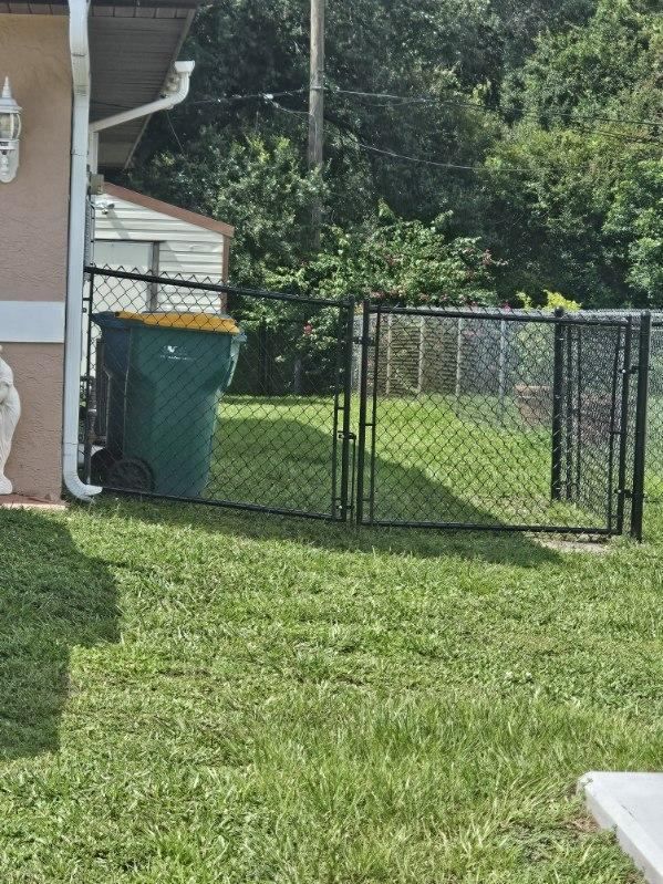 Backyard with chain-link dog pen, green trash bin, and grass beside a house
