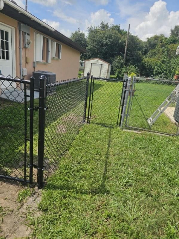 Black chain-link fence beside a tan house, opening to a grassy yard under a cloudy sky.