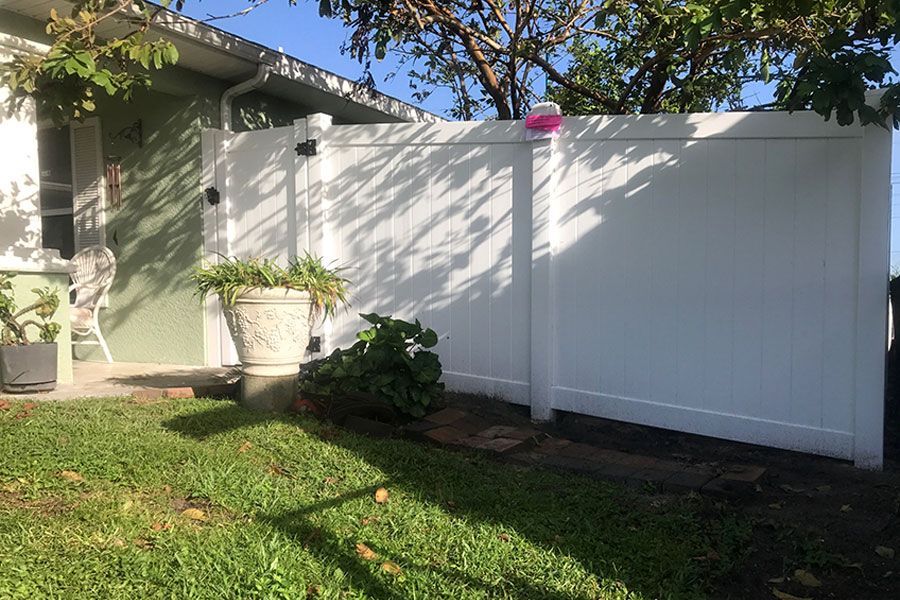 White fence beside a house with a potted plant and green lawn in a sunny yard.
