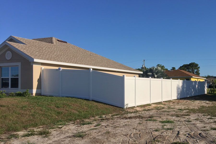 Beige house with white vinyl fence along a dirt lot under a clear blue sky