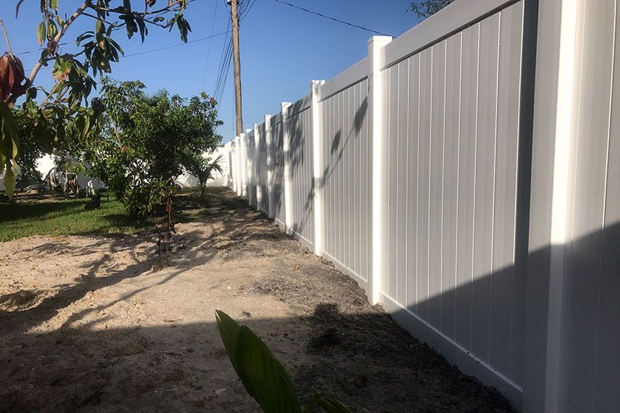 Sunlit dirt path beside a white vinyl fence and trees in a yard