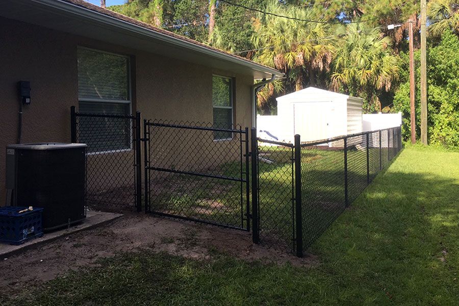 Black chain-link fence beside a beige house, bordering a grassy yard with a white shed in back