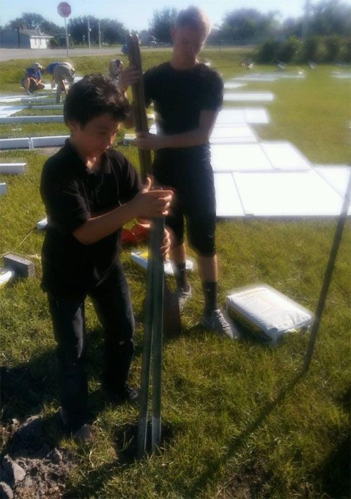 Two people in black shirts set up a metal tent frame on a grassy field with white pads in the background