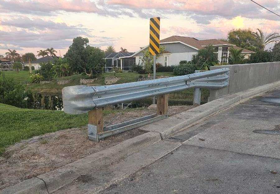 Roadside guardrail beside a cracked sidewalk at dusk, with houses and a striped warning sign in the background