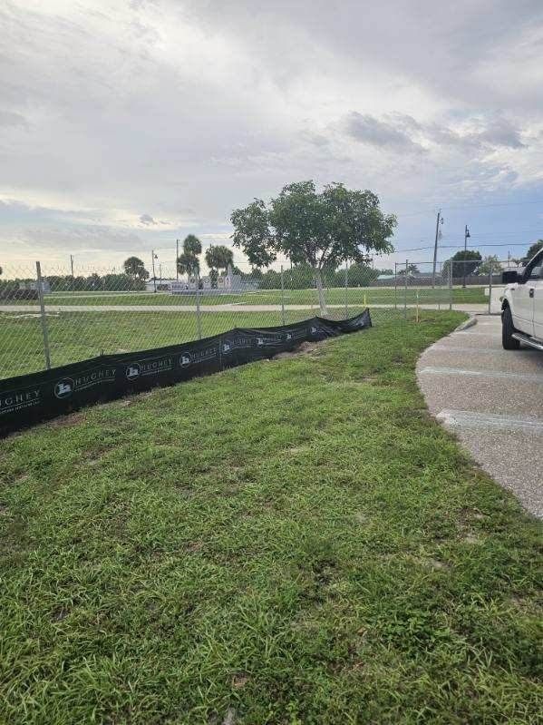 Grassy field beside a paved path, with a black barrier, fence, trees, and cloudy sky.