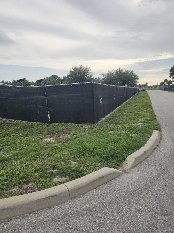 Black construction fence along a grassy roadside beside a curved asphalt street under a cloudy sky