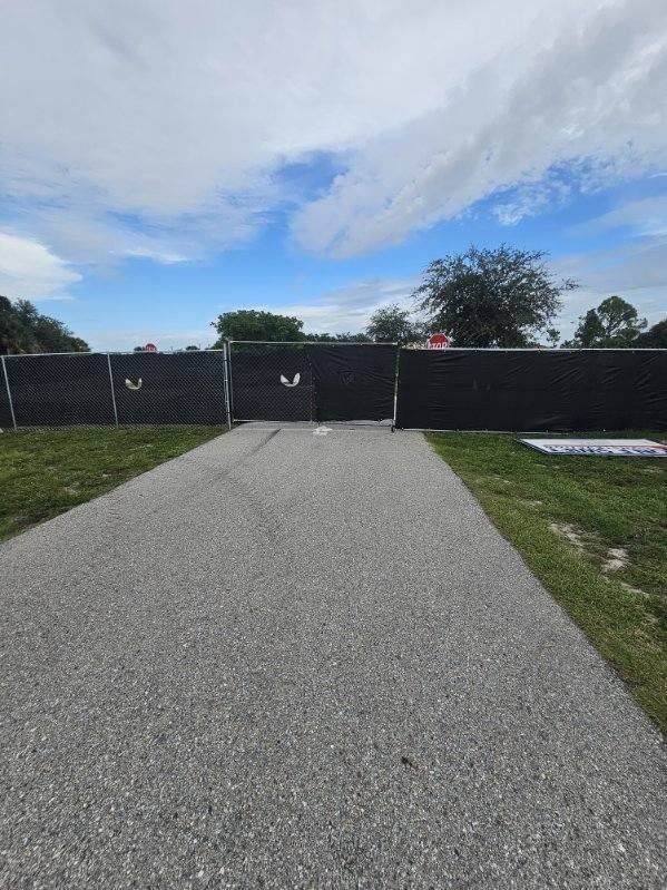 Gravel driveway leading to a black fence under a partly cloudy sky, with grass on both sides
