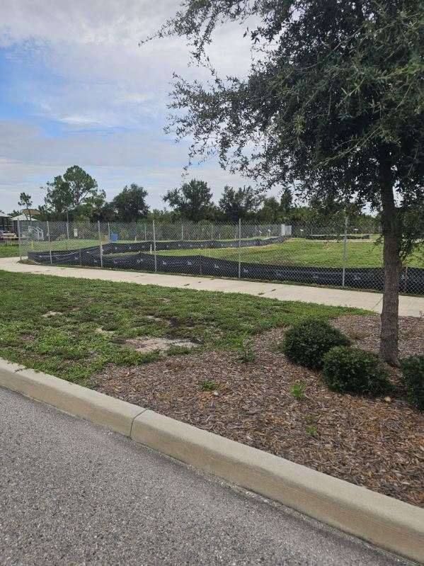 Sidewalk beside a landscaped area with a fenced field, trees, and overcast sky.