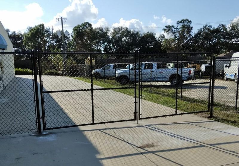 Black chain-link gate across a driveway with white pickup trucks and trees in the background