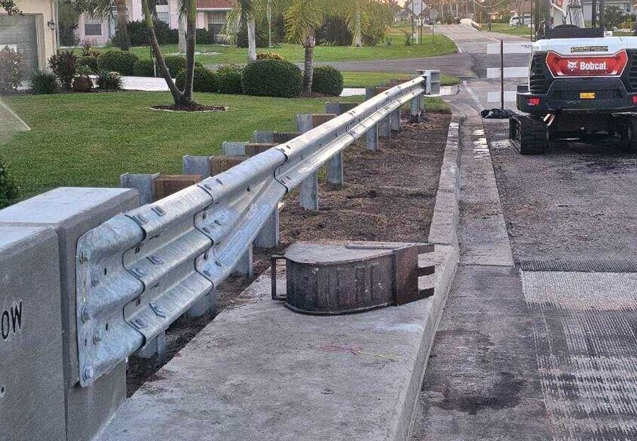 Damaged roadside guardrail beside a sidewalk, with a fallen post and a parked black car nearby.