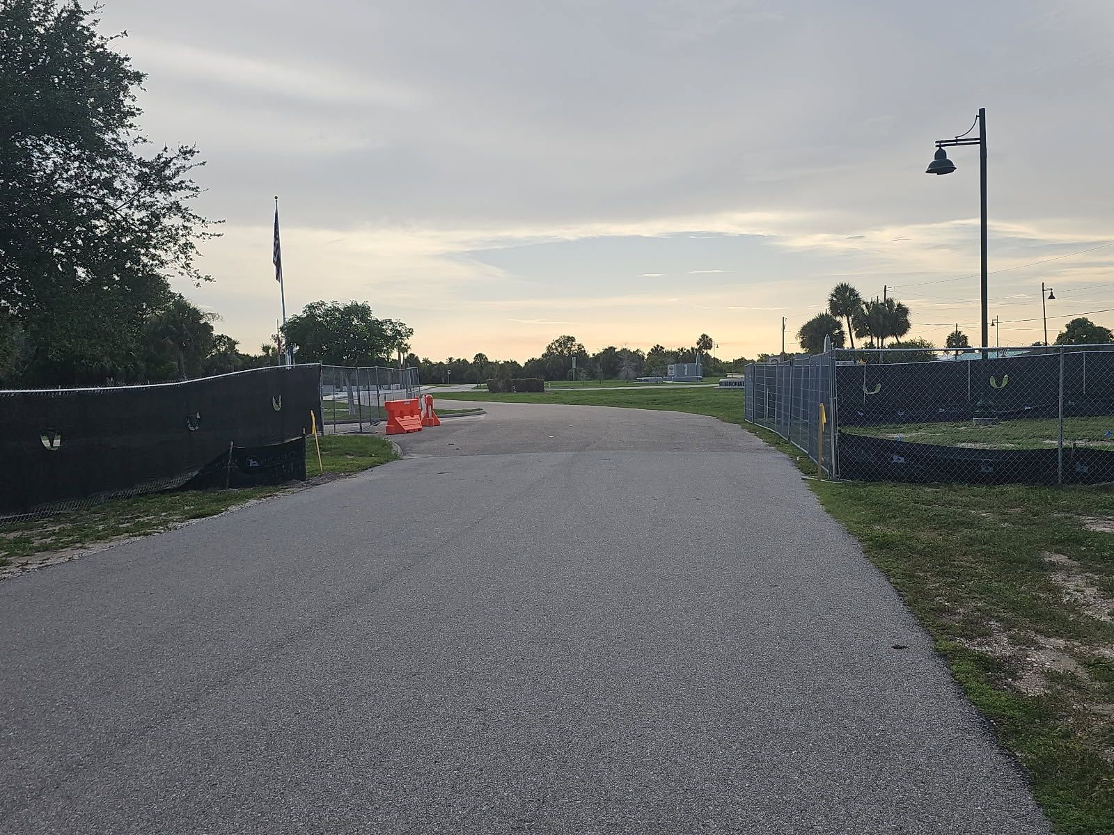 Gravel path between fenced areas at dusk, with orange barriers and a lamp post under a cloudy sky