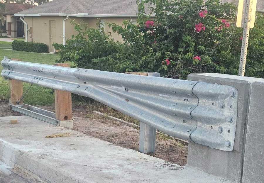 Damaged metal guardrail bent along a roadside concrete barrier near a residential area