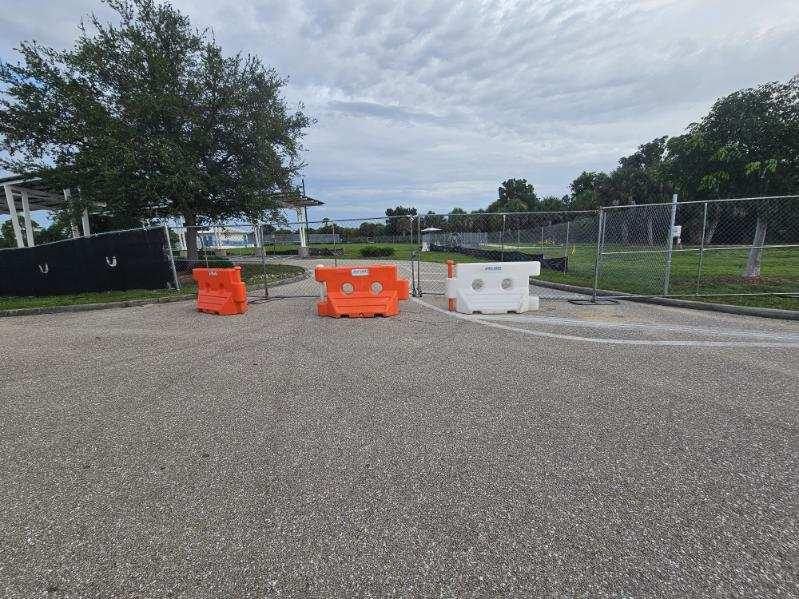 Orange and white barricades on a gravel lot near trees and a chain-link fence