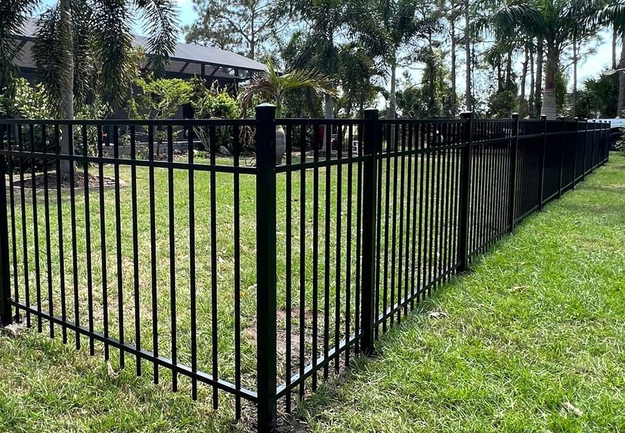Black metal fence enclosing a grassy yard with trees and a house in the background