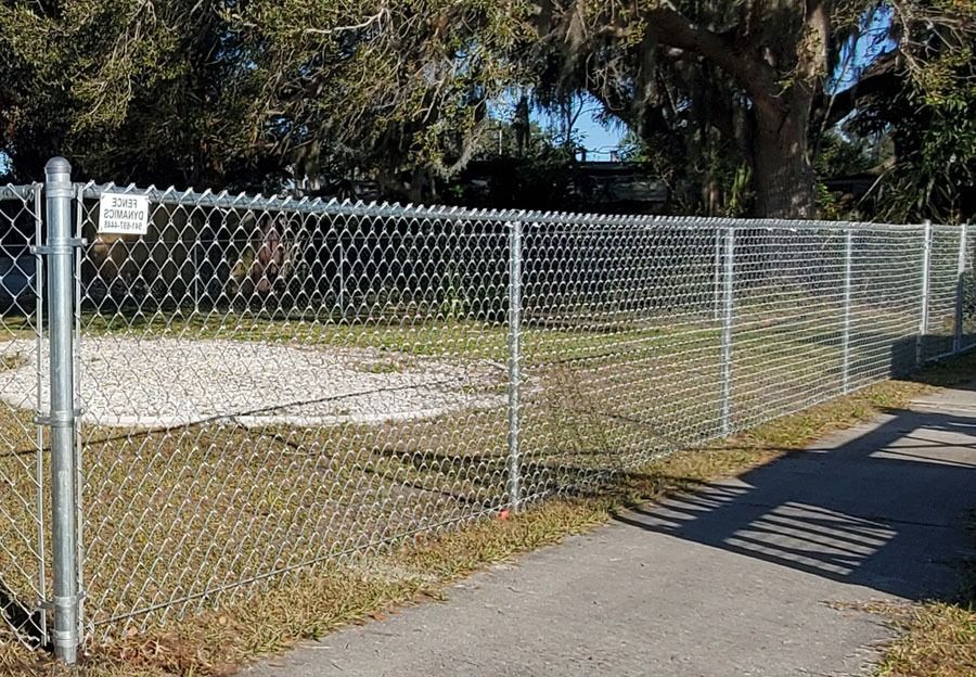 Chain-link fence beside a paved path and gravel lot under trees in bright sunlight