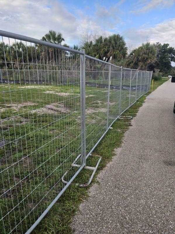 Temporary metal fence along a paved path beside grass and trees under a partly cloudy sky