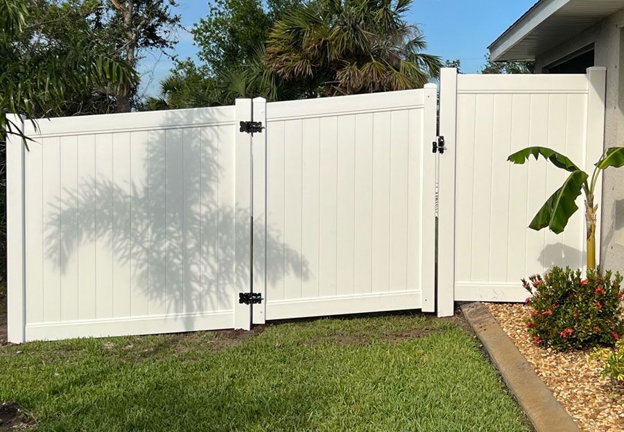 White vinyl backyard fence with gate beside a lawn and tropical plants