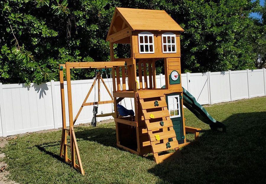 Wooden backyard playset with slide, swing, ladder, and tower beside a white fence on grass.