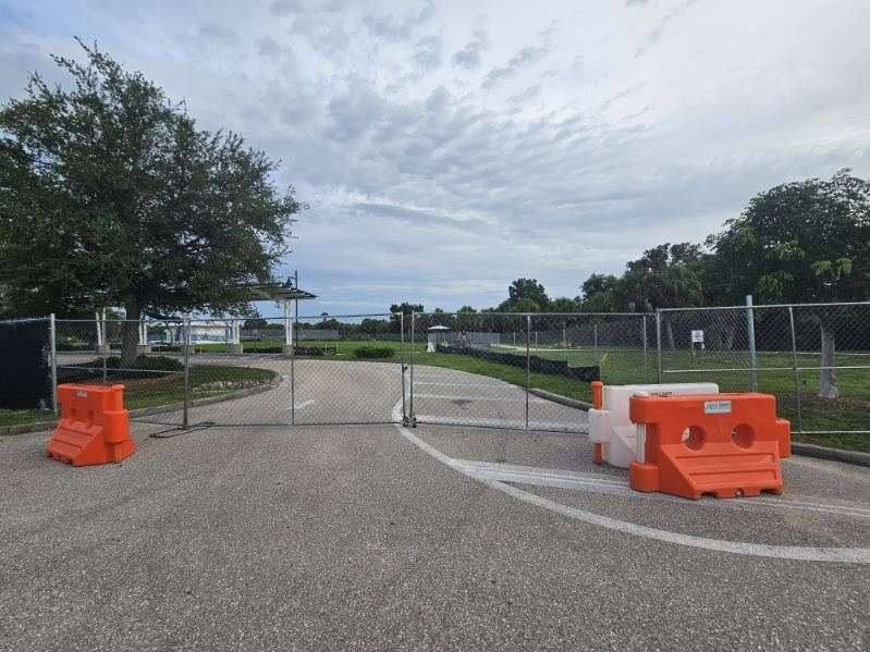 Blocked paved path with orange barricades and a gate, leading into a tree-lined park under cloudy sky