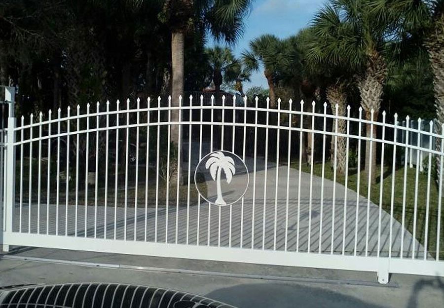White metal driveway gate with a circular emblem in front of a palm-lined property