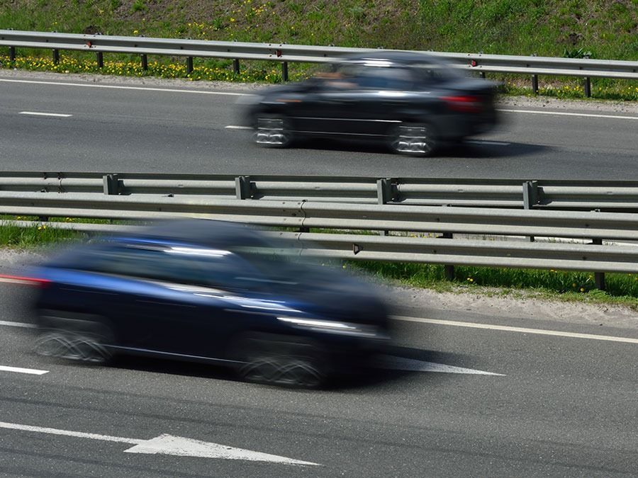 Two blurred cars speeding on a highway with metal guardrails and greenery in the background