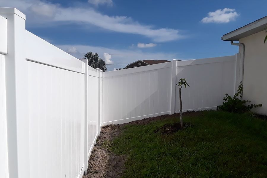 White vinyl fence lining a narrow grassy backyard under a blue sky with clouds.