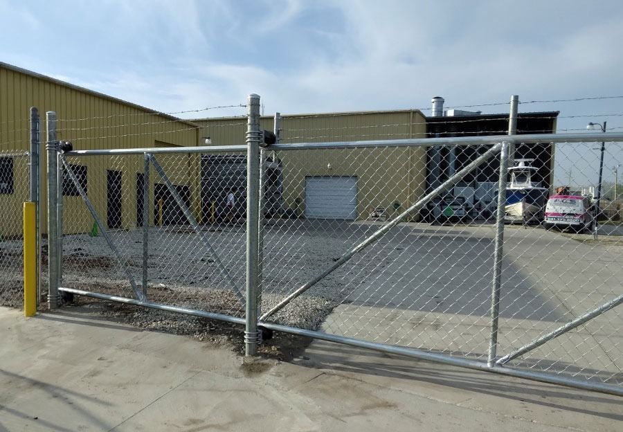 Chain-link gate in front of an industrial warehouse and parking lot, with a parked car in the background.