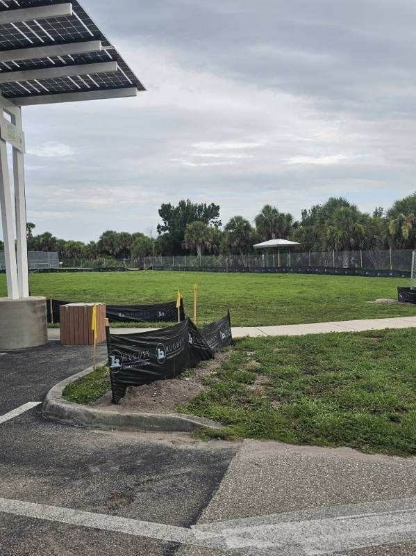 Outdoor park path beside a grassy field, with a black mesh barrier and cloudy sky.