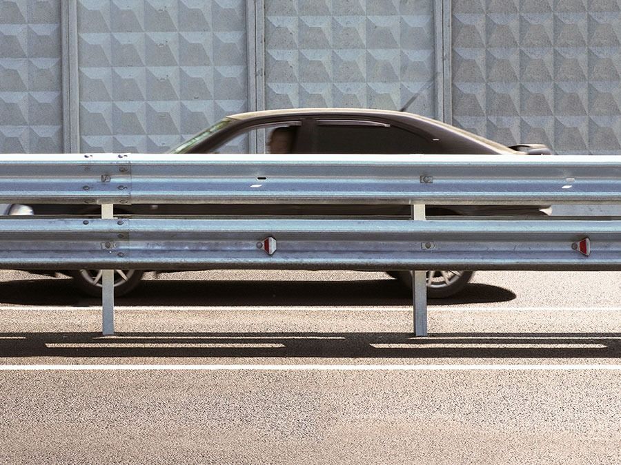 Silver sports car behind a blue guardrail on a roadside, viewed from the side.