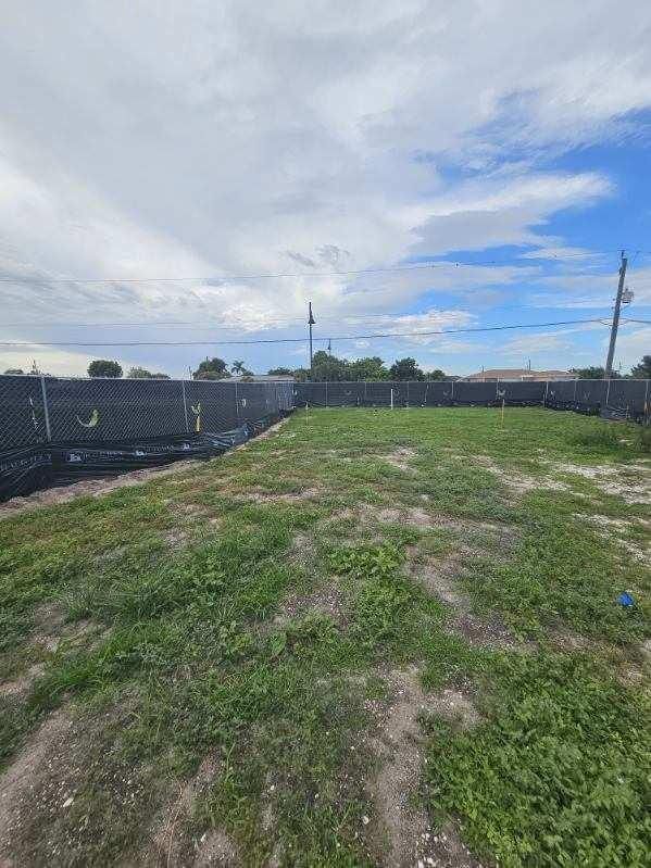 Overgrown grassy lot with a dirt path, chain-link fence, and cloudy sky