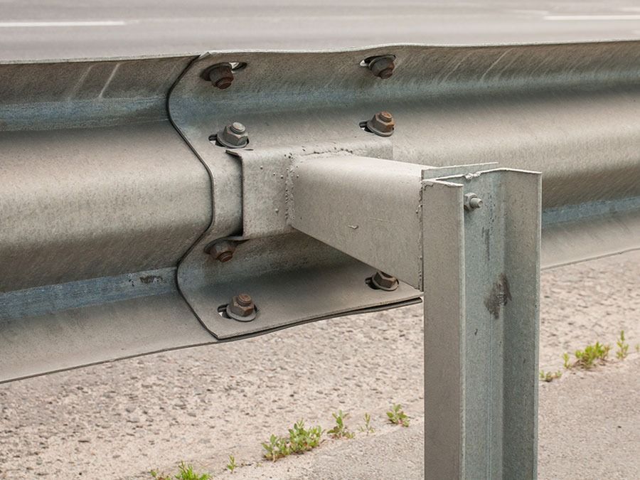Close-up of a metal guardrail support bolted to a silver roadside barrier on pavement.