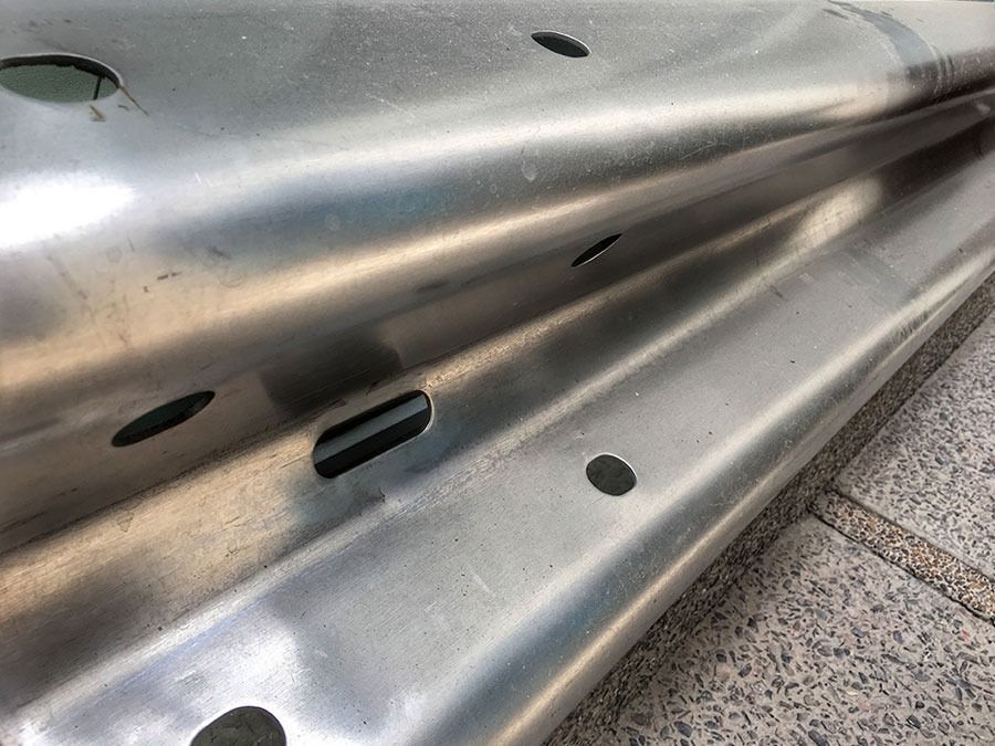 Close-up of a shiny metal guardrail with slotted holes and a concrete sidewalk beside it