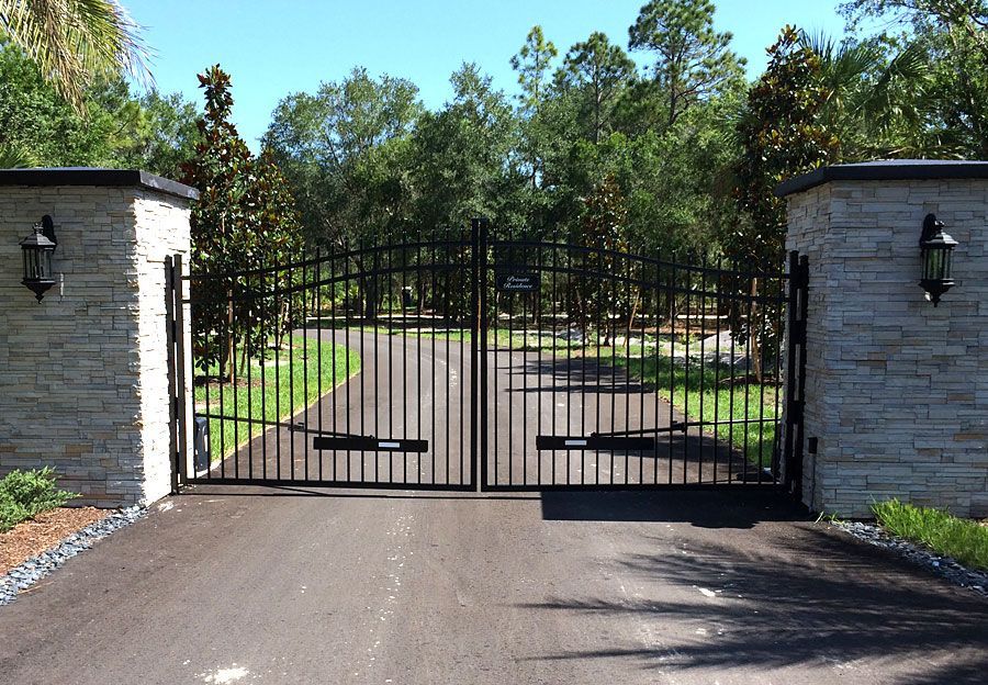 Black metal gate between stone pillars at a driveway entrance, with trees and a paved road beyond.