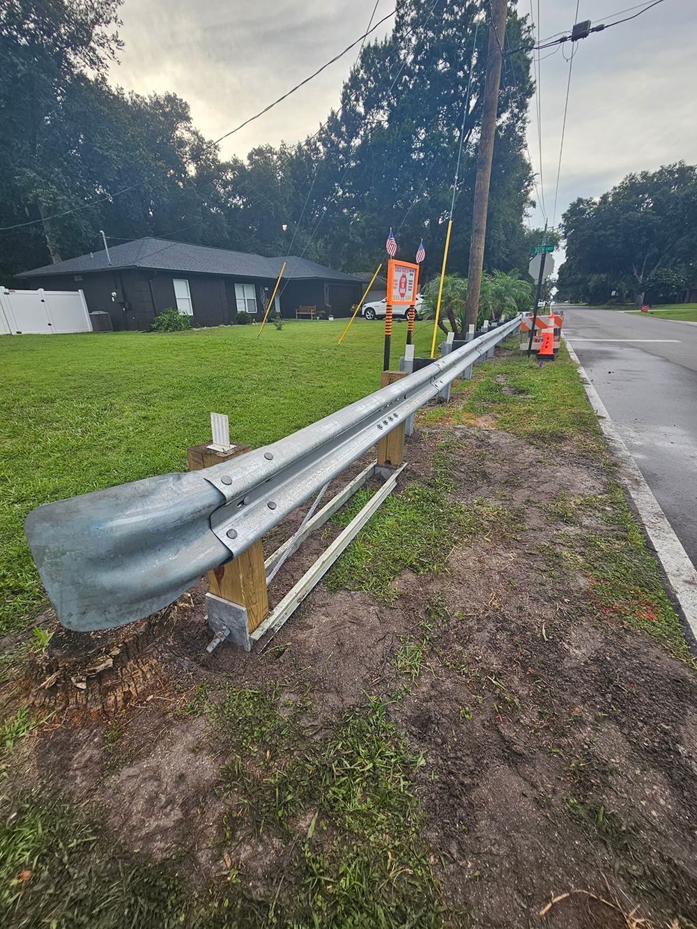 Damaged roadside guardrail beside a wet street, with cones and a worker in the distance.