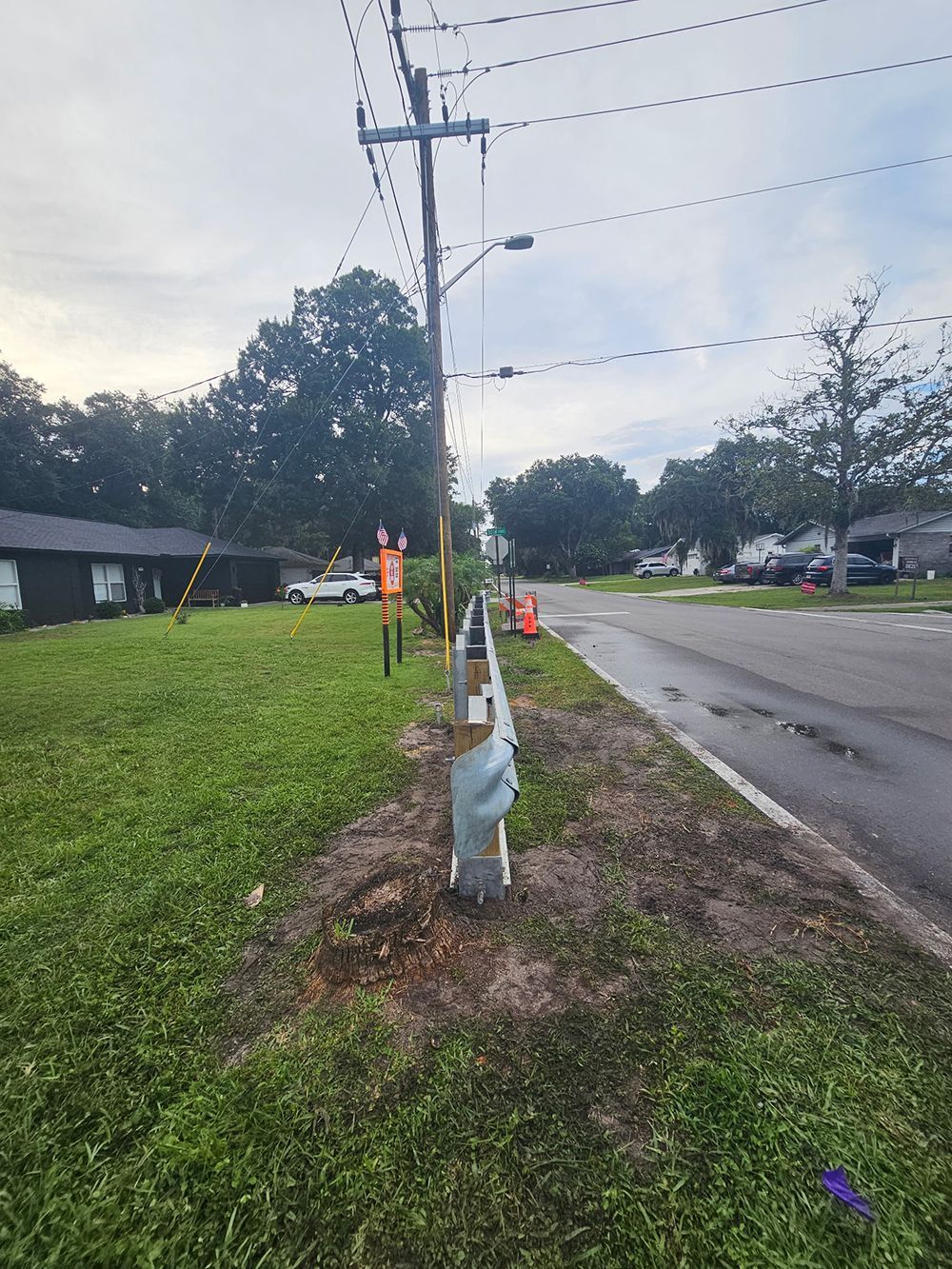 Utility pole beside a wet roadside with grassy verge, cloudy sky, and houses in the background.